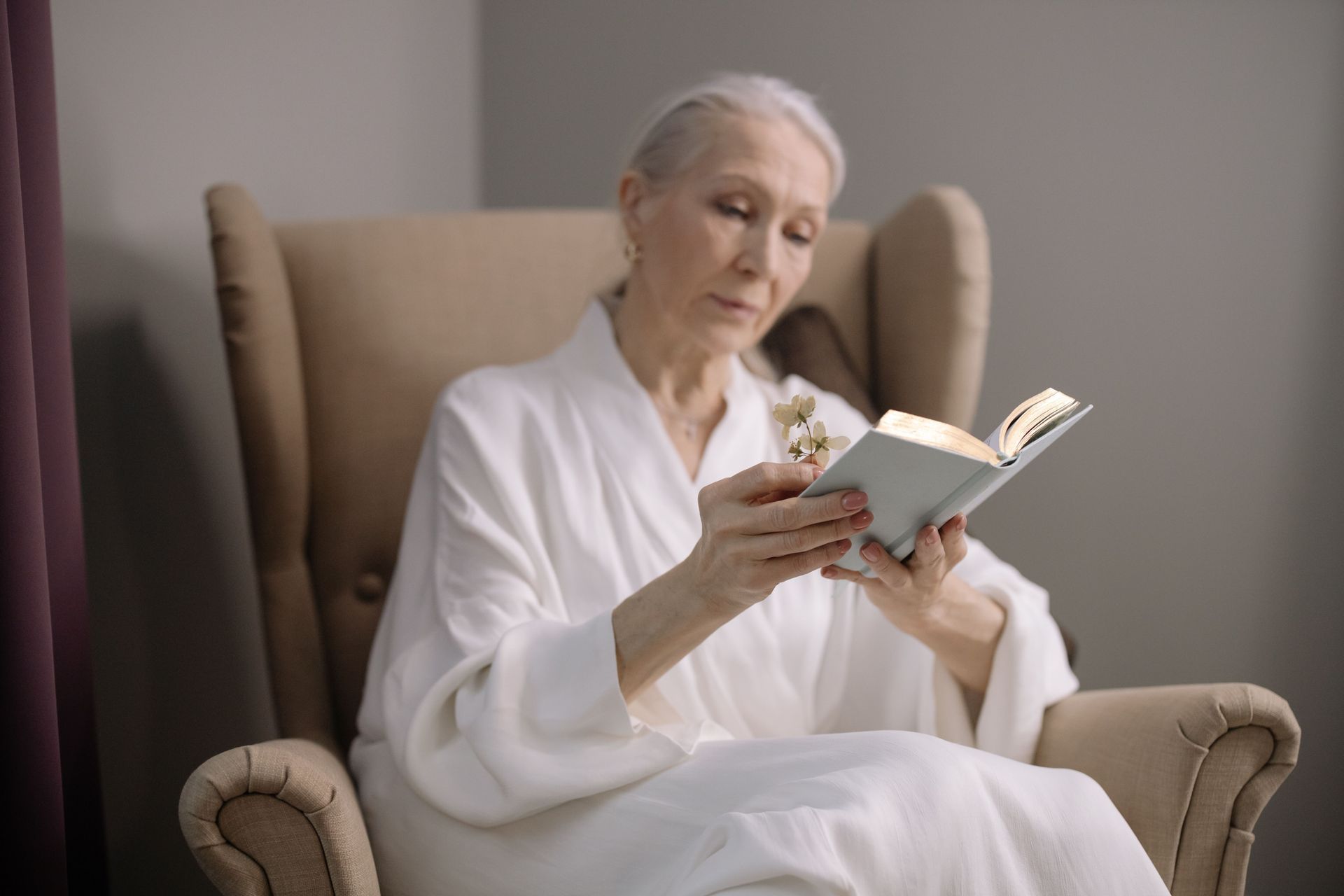 Person in a white robe reading a book while seated in a beige armchair