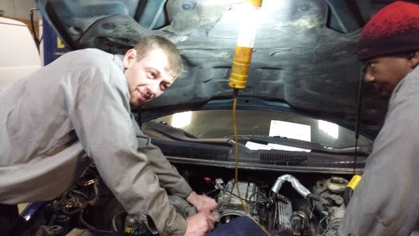 Two mechanics working under a car hood, illuminated by a hanging work light.