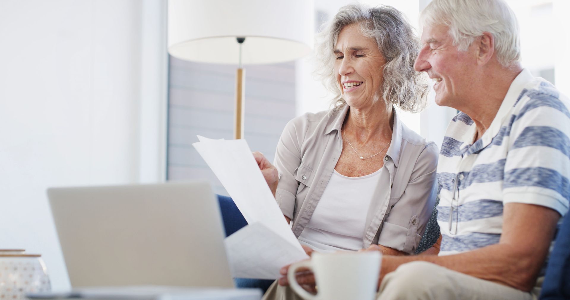 Two smiling adults reviewing papers beside a laptop in a bright living room