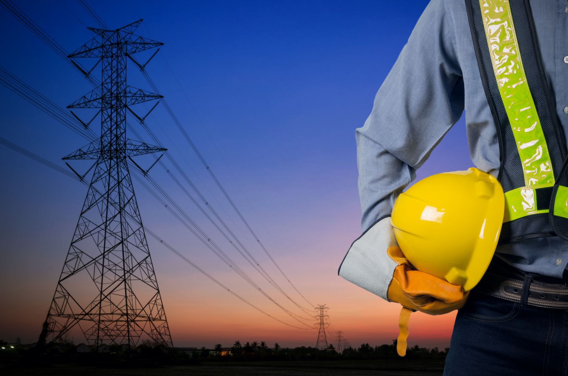 Electrician holding a yellow hard hat beside power lines at dusk