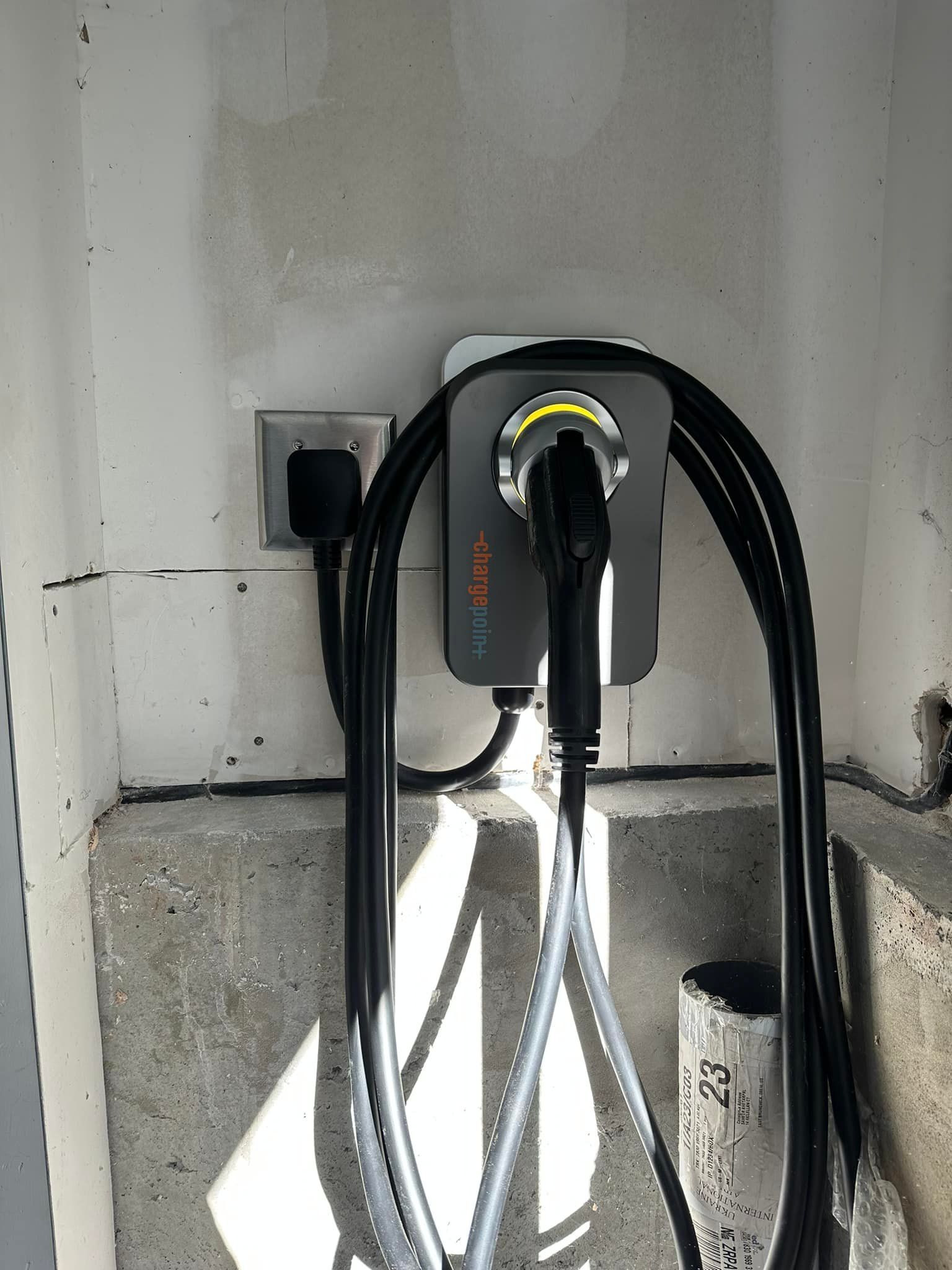 Wall-mounted EV charger with coiled black cable in a white garage beside an outlet.