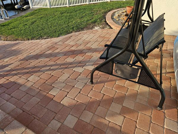 Brick patio with a black metal swing seat. Green grass and white fence in background.