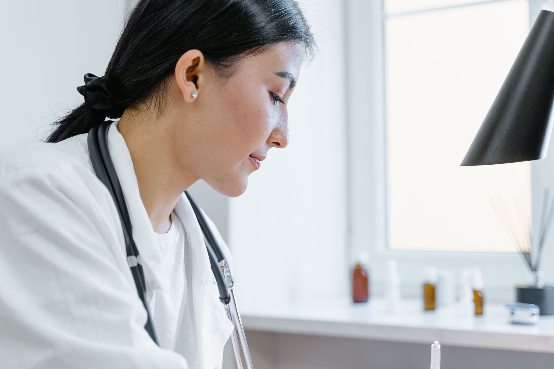 Doctor in a white coat writing at a desk by a bright window