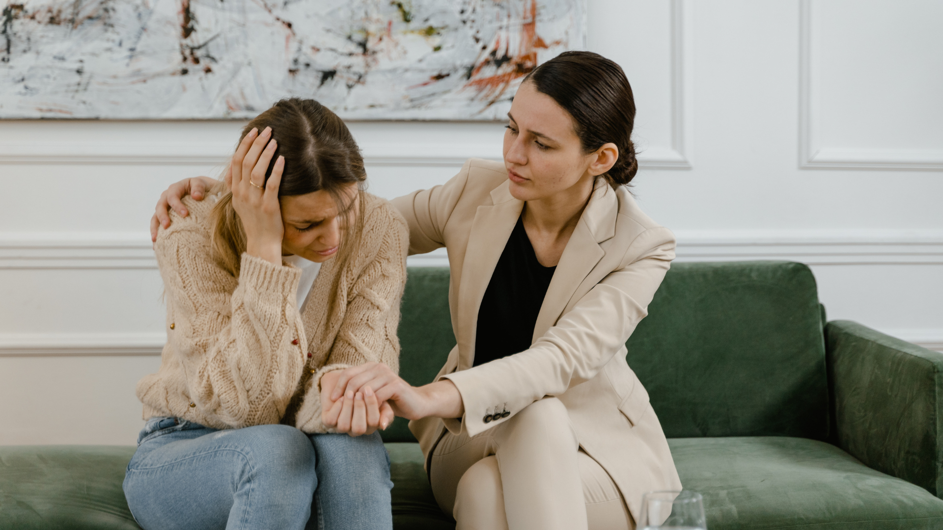 Supportive woman comforts a distressed person sitting on a couch in a bright living room.