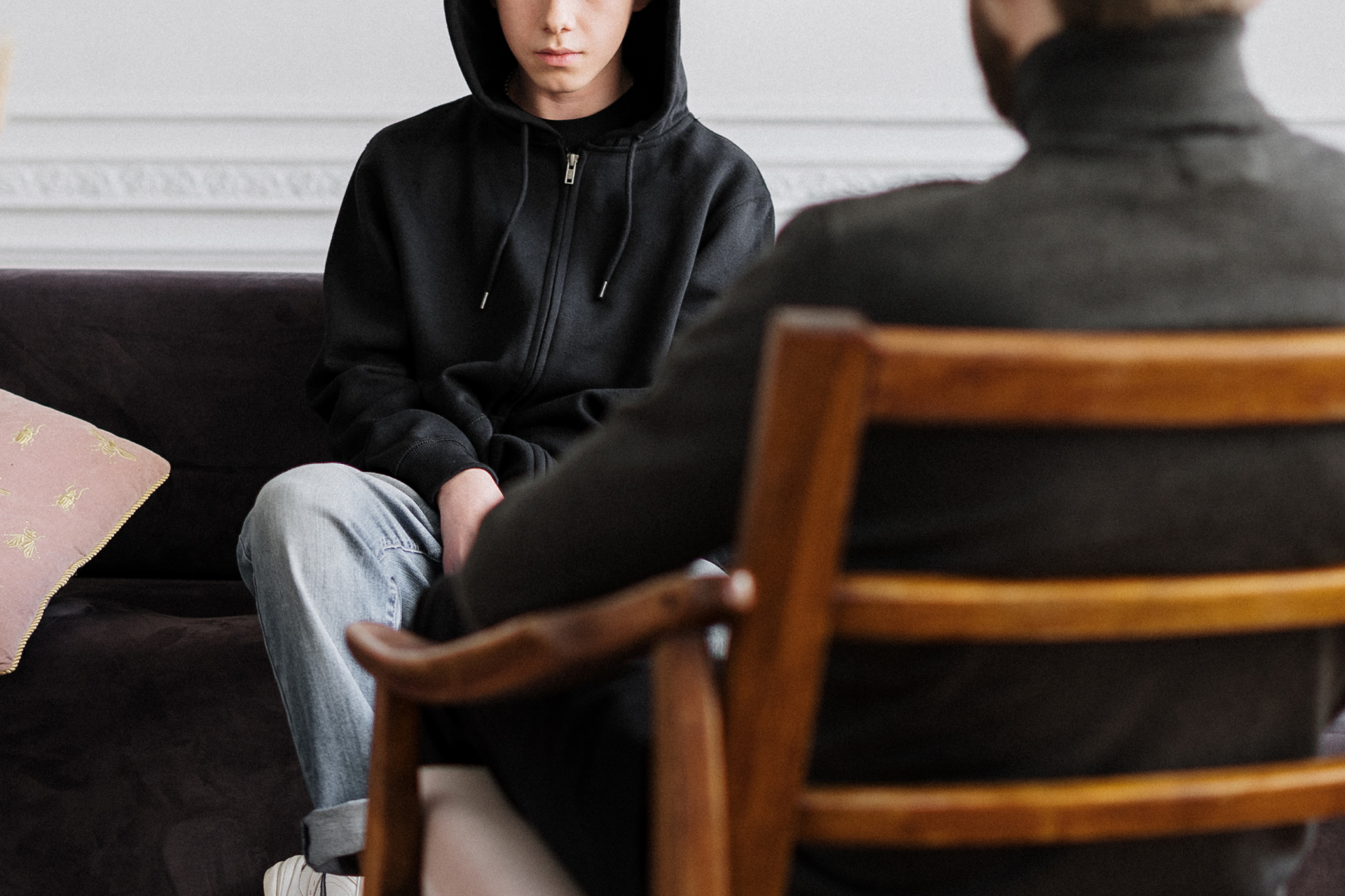 Two people seated in a living room, one in a black hoodie facing the other in a wooden chair.