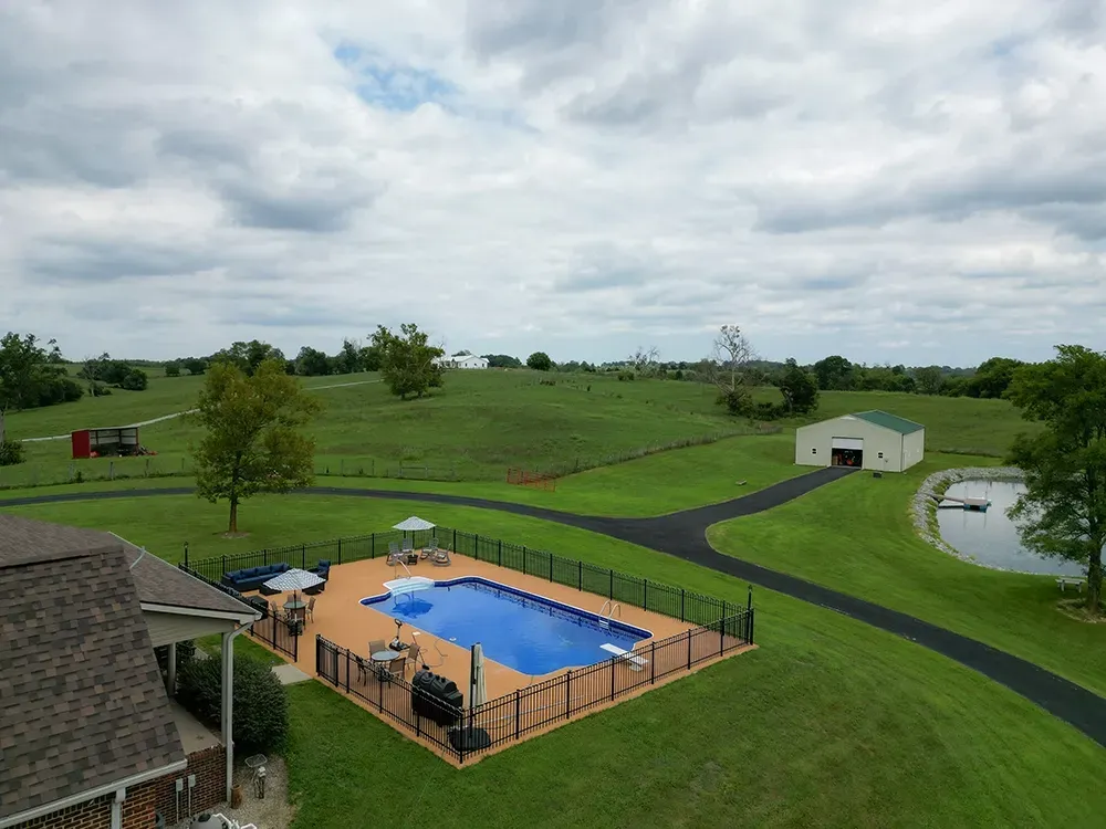Swimming pool and property with green grass, trees, and buildings under a cloudy sky.