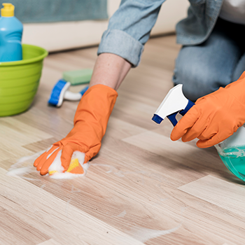 Person cleaning a wooden floor with orange gloves and spray cleaner near a bucket and brush