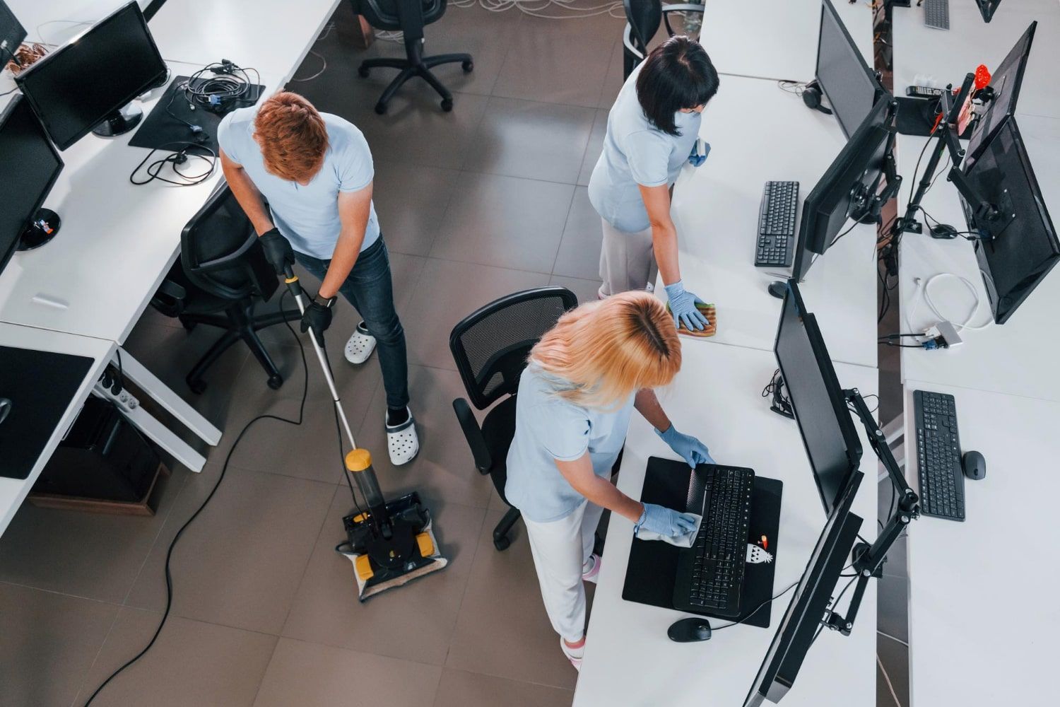 Three people cleaning a modern office workspace with computers and desks.