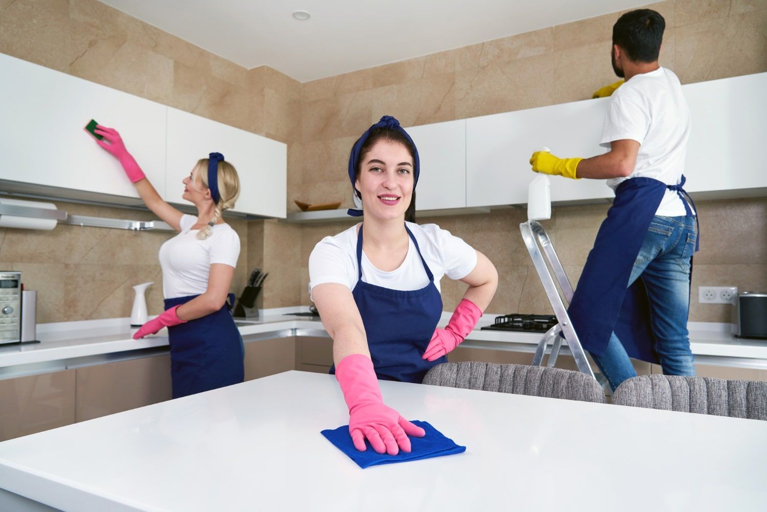 Three cleaners in a bright kitchen, wearing aprons and pink gloves, wiping counters and shelves.