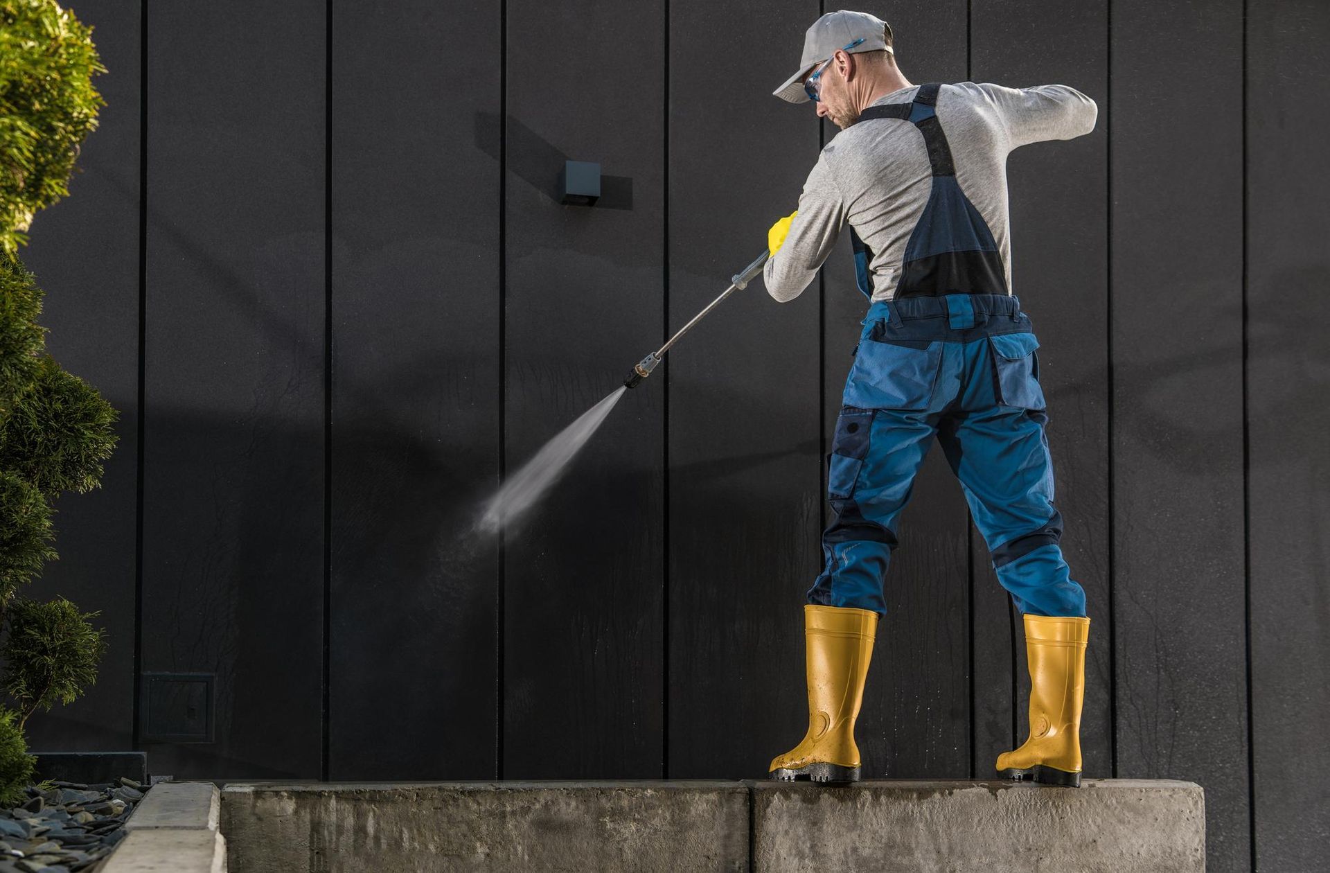 Worker in blue coveralls and yellow boots pressure washing a dark wall outdoors.
