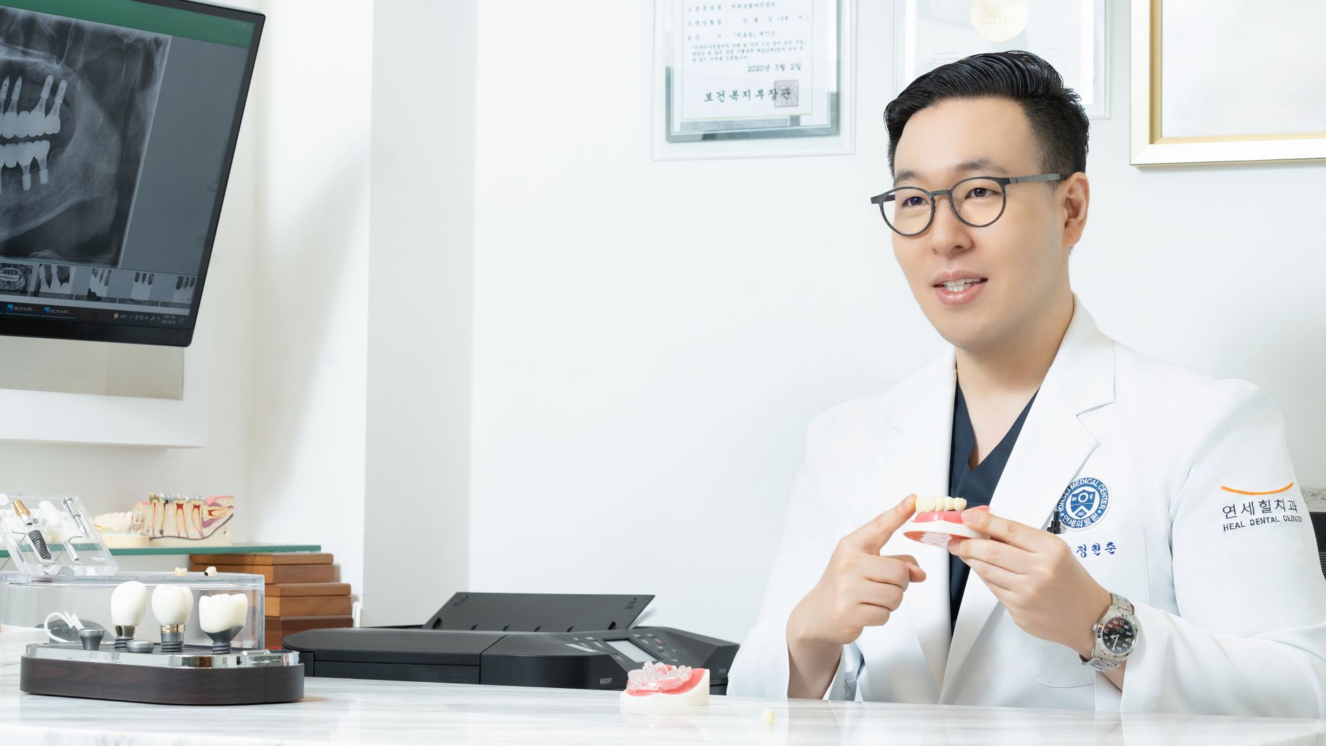 A smiling dentist in a white coat gestures with dental models in a bright office with an x-ray.