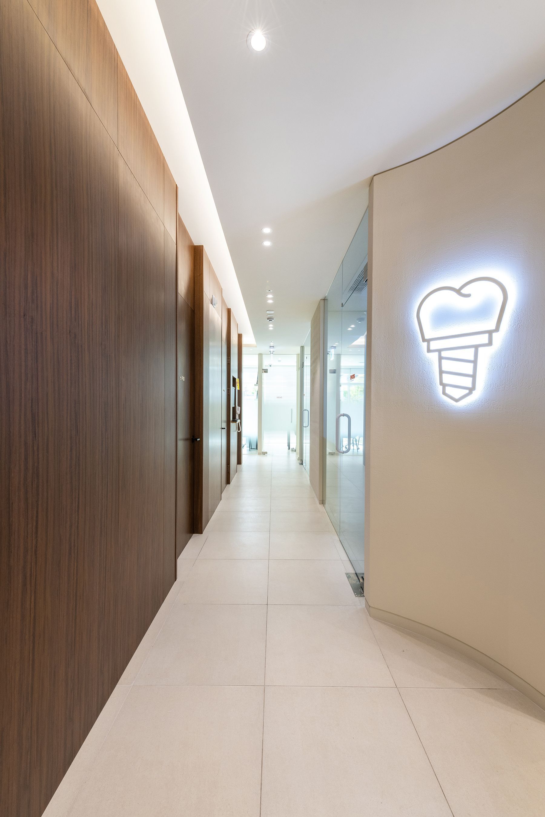 A brightly lit dental office hallway with wooden paneling, doors, and a lit tooth logo.