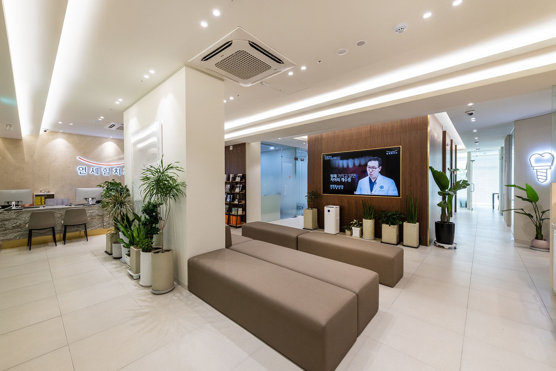 Modern waiting room with brown benches, plants, and a TV screen.