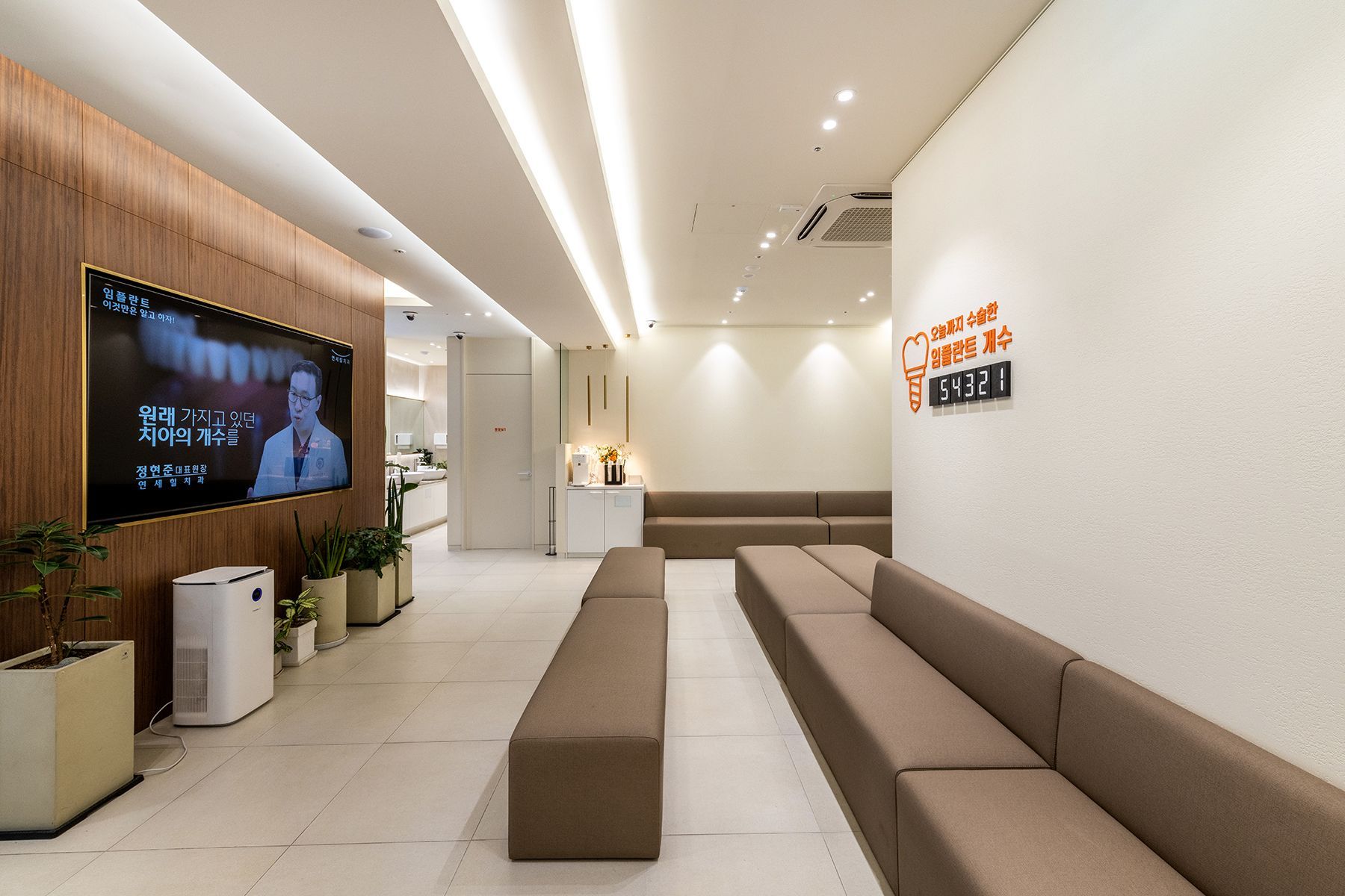 Modern dental clinic waiting room; brown benches, TV, white walls, orange sign.