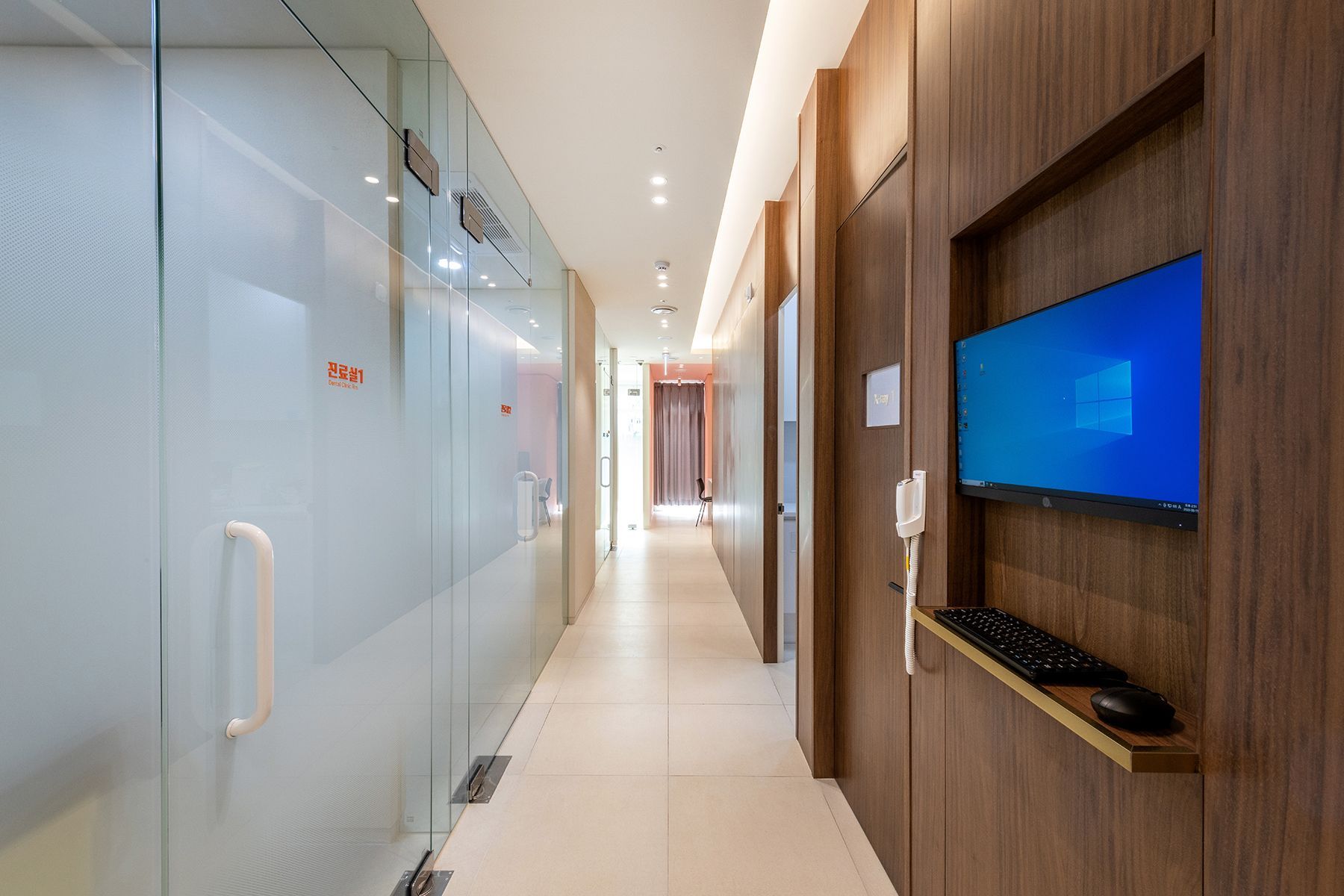 Long hallway with glass walls, wood paneling, a computer, and doors in a modern office.
