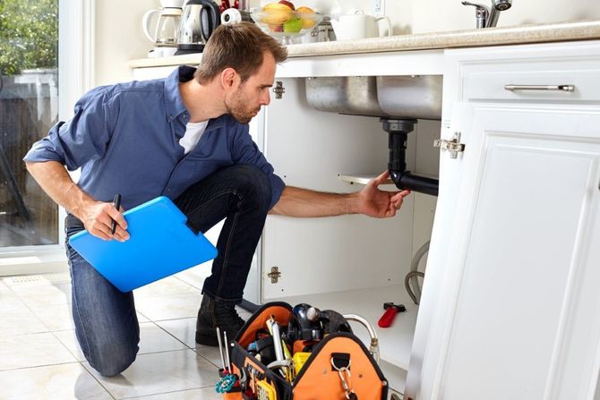 Technician inspecting plumbing under a kitchen sink with tools nearby