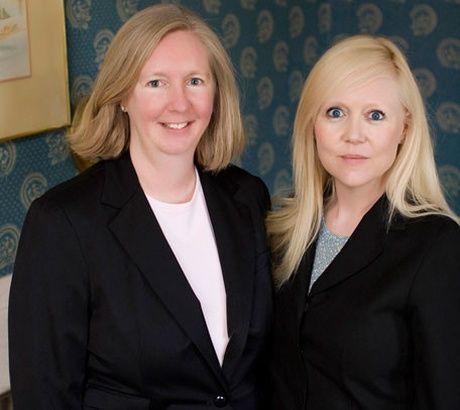 Two women in black blazers standing indoors, smiling at the camera.
