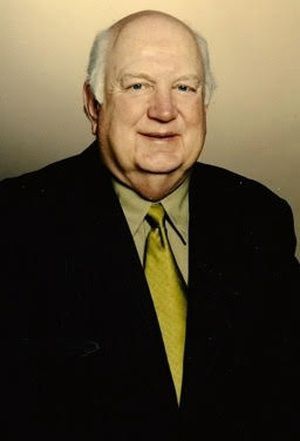 Portrait of a man in a dark suit and yellow tie against a plain beige background