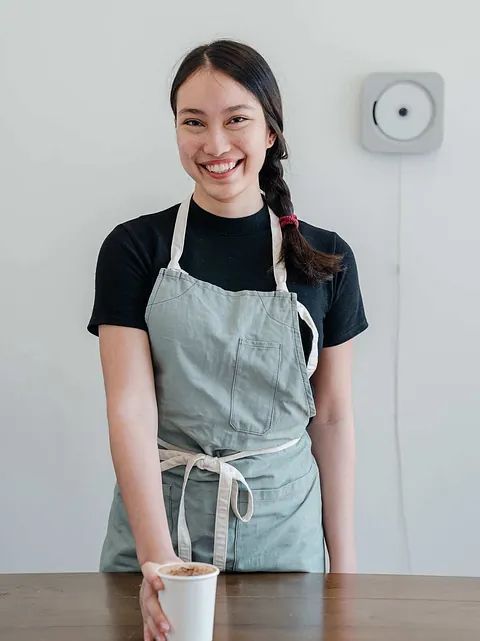 Smiling person in a gray apron stands behind a table with a cup of coffee in a bright room.