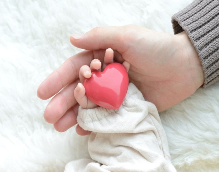 Hand holding a small pink heart in a baby’s clenched fist on a white blanket