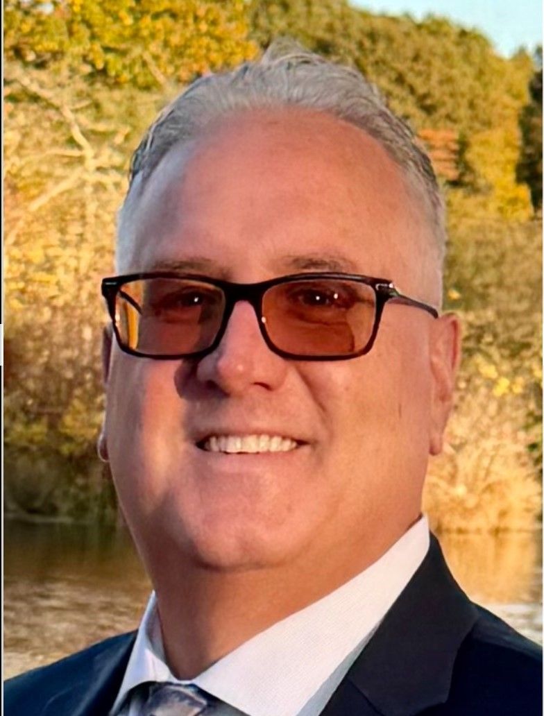 Man in glasses smiling, wearing suit jacket and tie, outdoors with fall foliage.