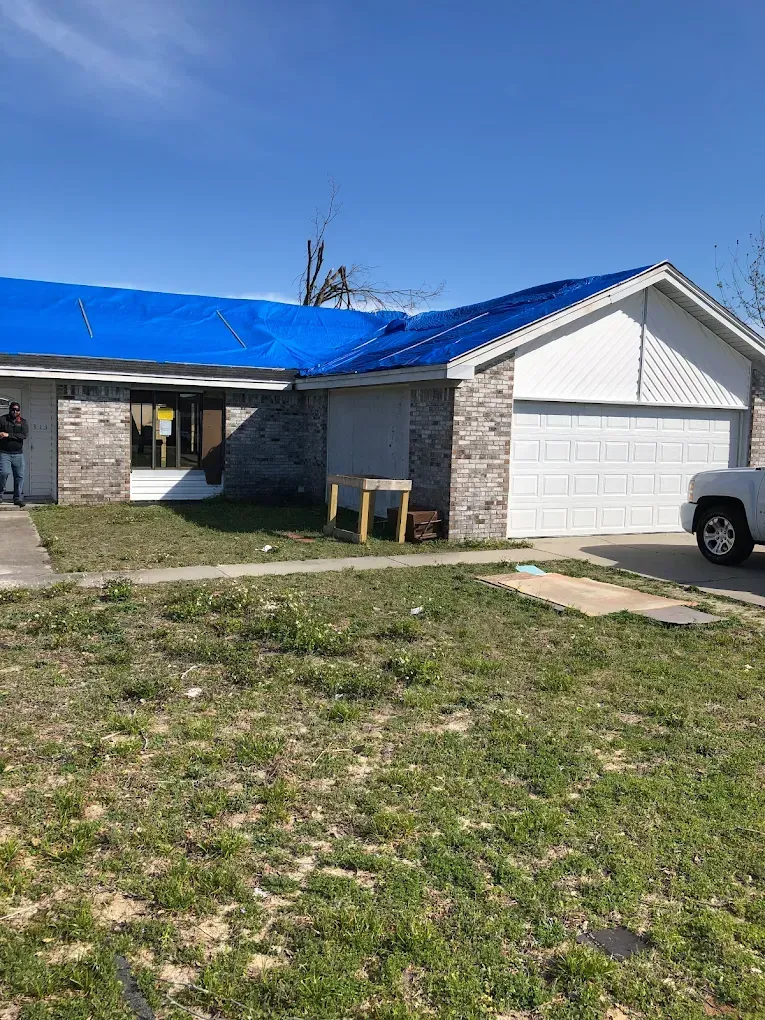 A house with a blue tarp covering the roof, indicating repair or storm damage, with a white garage and front yard.