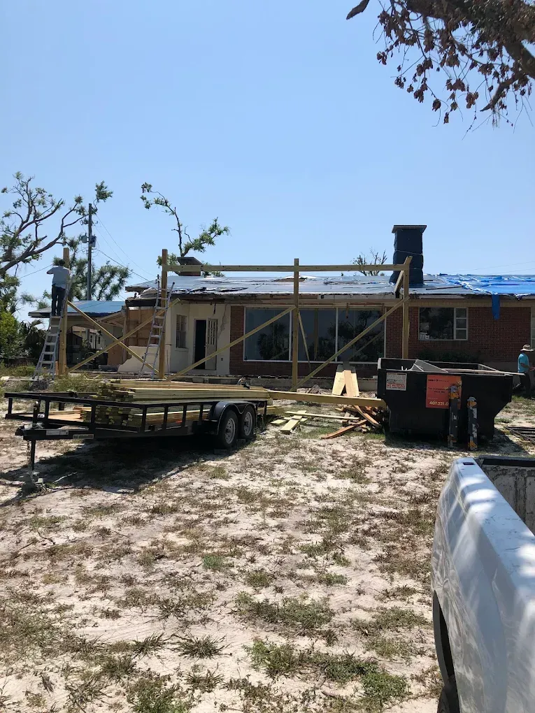 A person stands on a ladder working on the wooden framing of a house exterior under repair, with a trailer and dumpster.