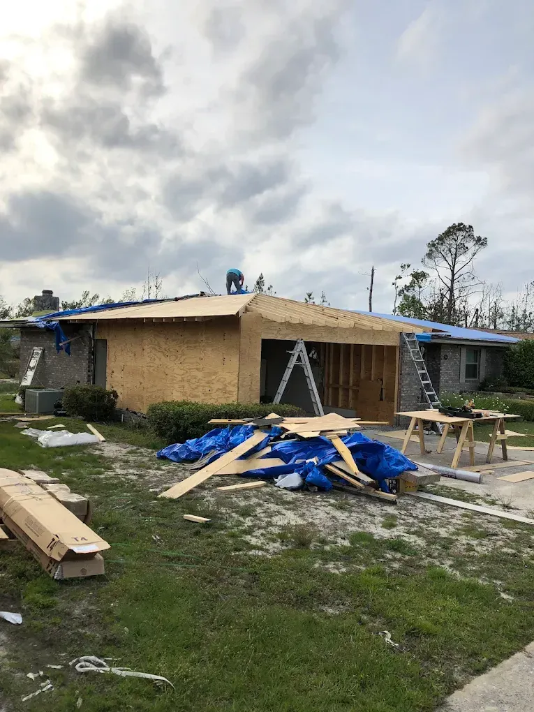 Construction workers repairing a damaged house roof and exterior with plywood and ladders under a cloudy sky.