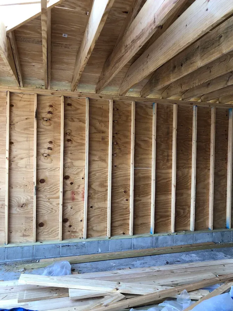 Unfinished wood-framed wall with plywood sheathing sitting on a concrete block foundation under exposed ceiling joists.