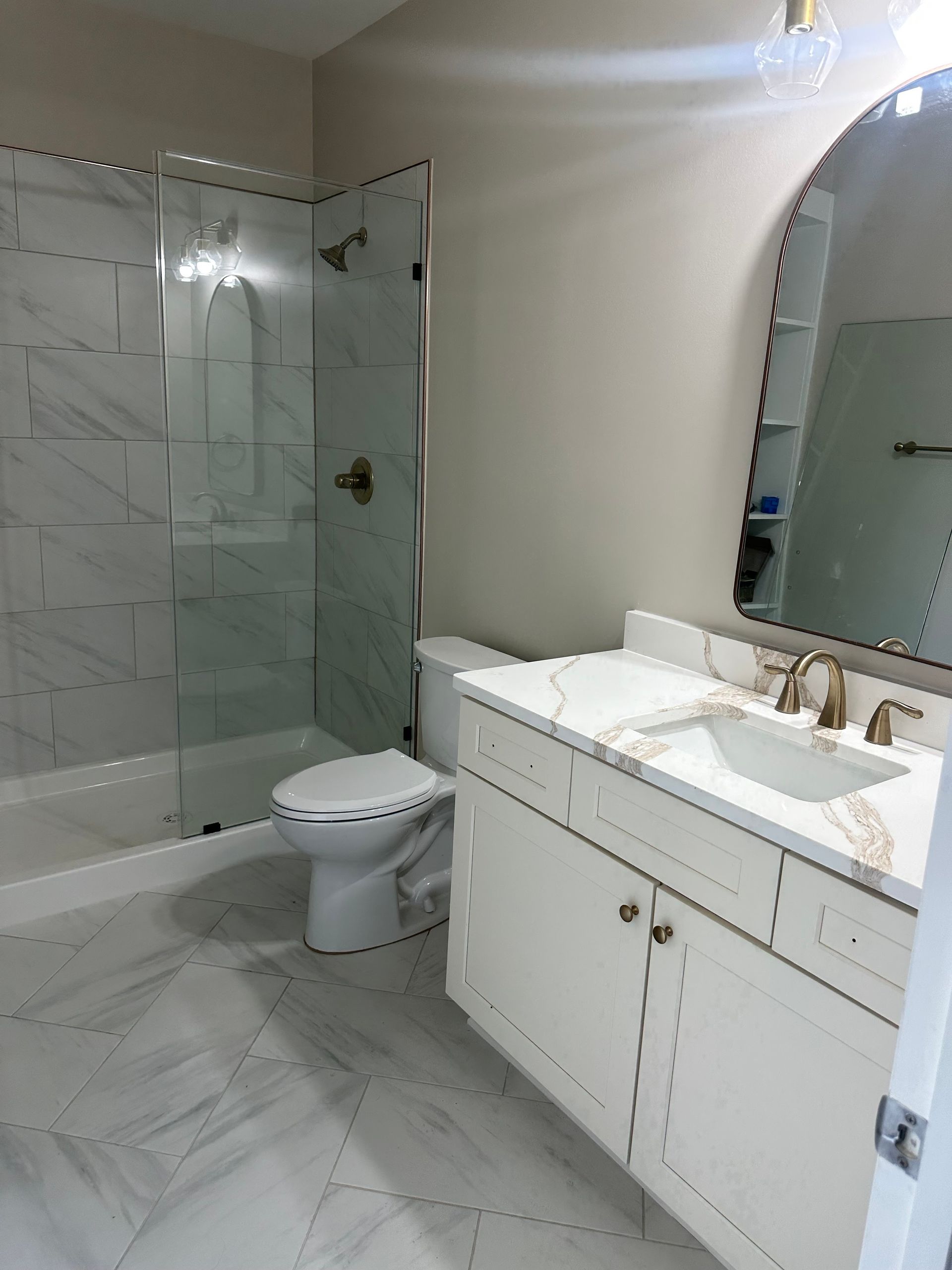 A bright bathroom with a marble-tiled shower, white vanity, toilet, and an arched mirror on a neutral wall.