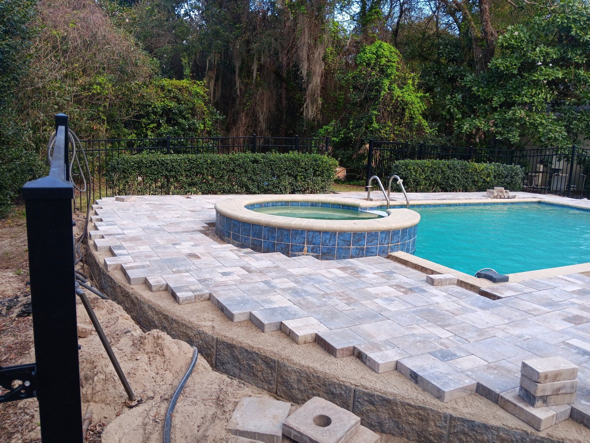 Newly laid stone patio pavers around a backyard swimming pool and hot tub, with an unfinished edge and dirt area.