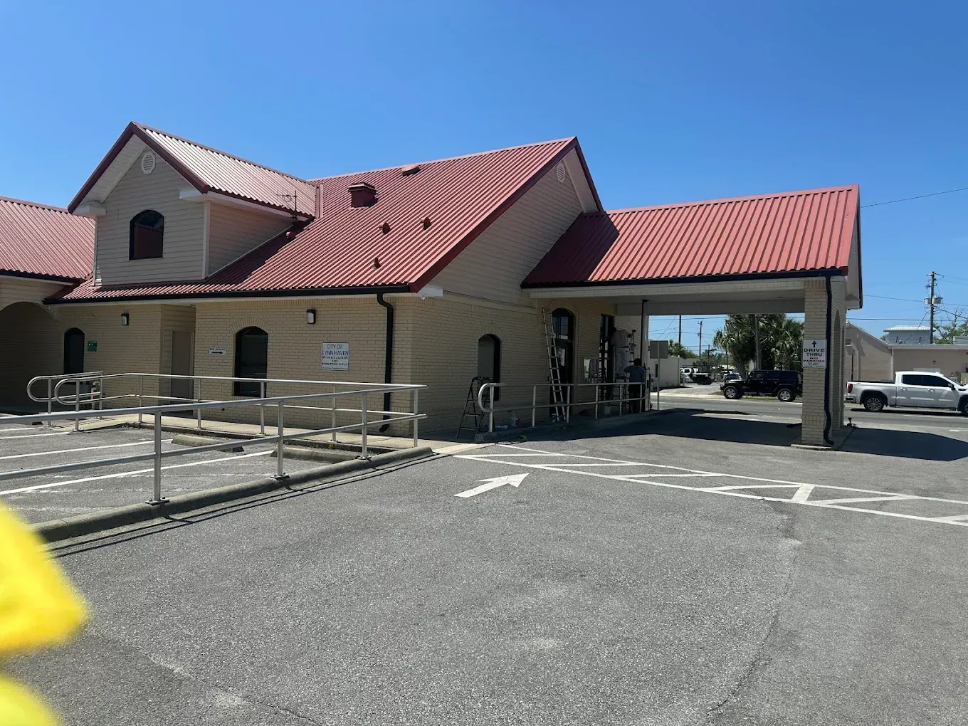 A brick building with a red roof, a drive-thru canopy, and a metal ramp entrance in a parking lot on a sunny day.