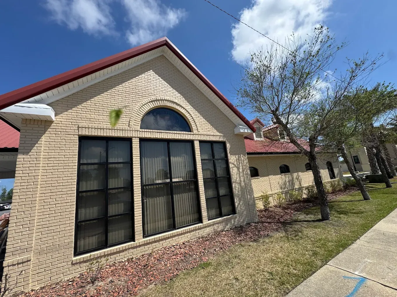 Tan brick building with a red roof, a large arched window, and a sidewalk leading past it under a clear blue sky.