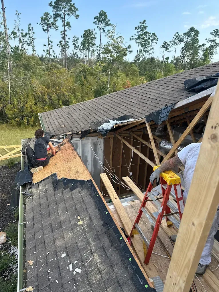 Two construction workers repair a damaged shingled roof on a sunny day, with one worker standing on a ladder.