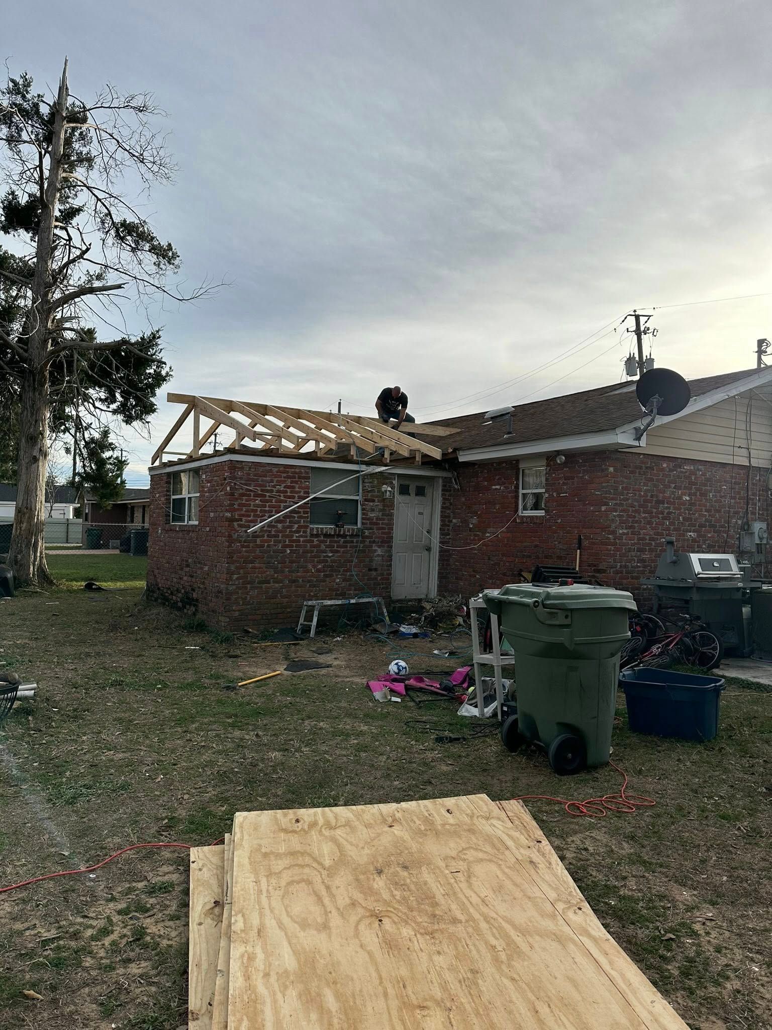 A brick house with a partially destroyed roof featuring exposed wooden trusses, with construction materials in the yard.