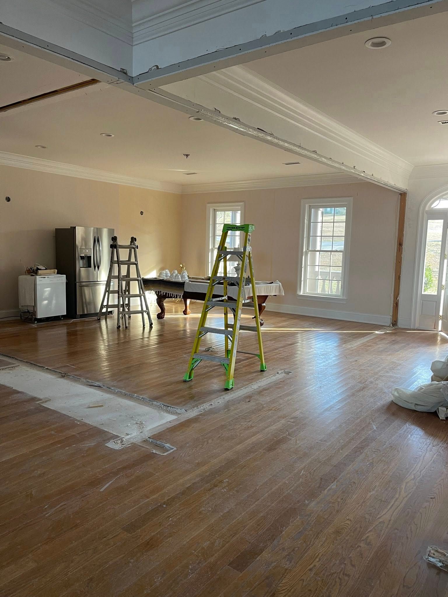 An indoor renovation scene shows a large, empty room with hardwood floors, a stepladder, a refrigerator, and molding.