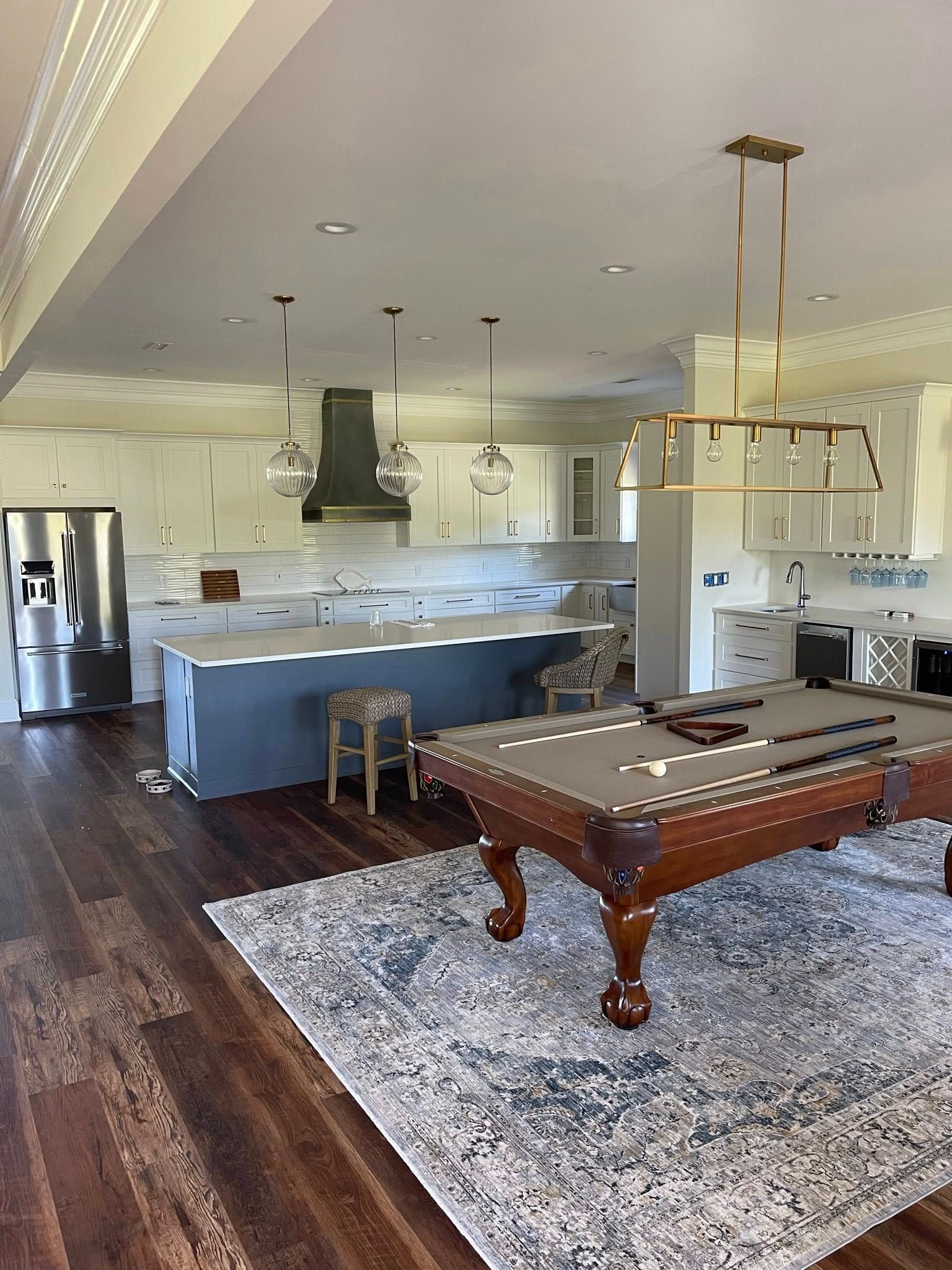A pool table with a patterned rug sits in front of a white kitchen with a grey island and gold pendant lighting.