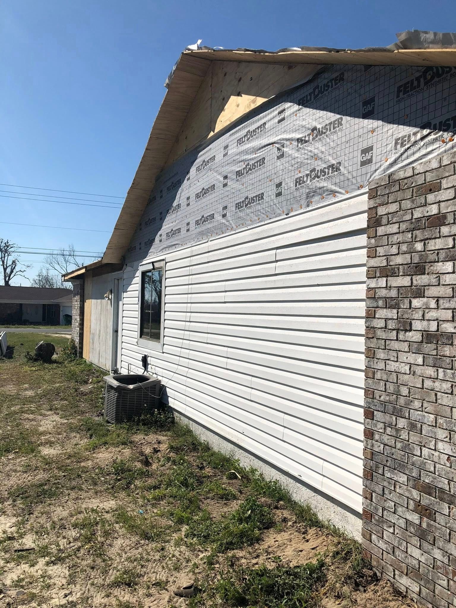 An exterior wall of a house under construction, featuring partial white siding, exposed gray house wrap, and brick veneer.