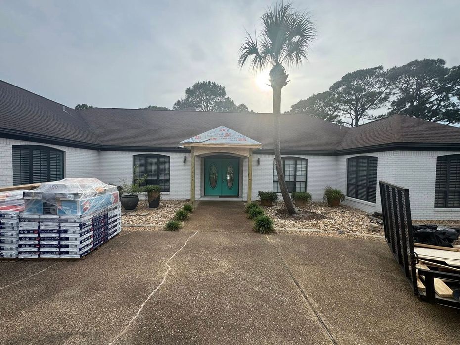 A white, single-story house with a teal front door, a palm tree in the front, and construction materials on the driveway.