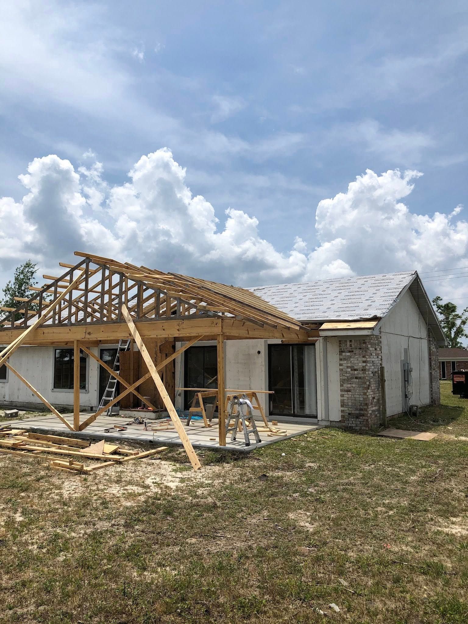 A house under construction with a wooden roof frame attached to a brick-and-stucco structure under a cloudy sky.