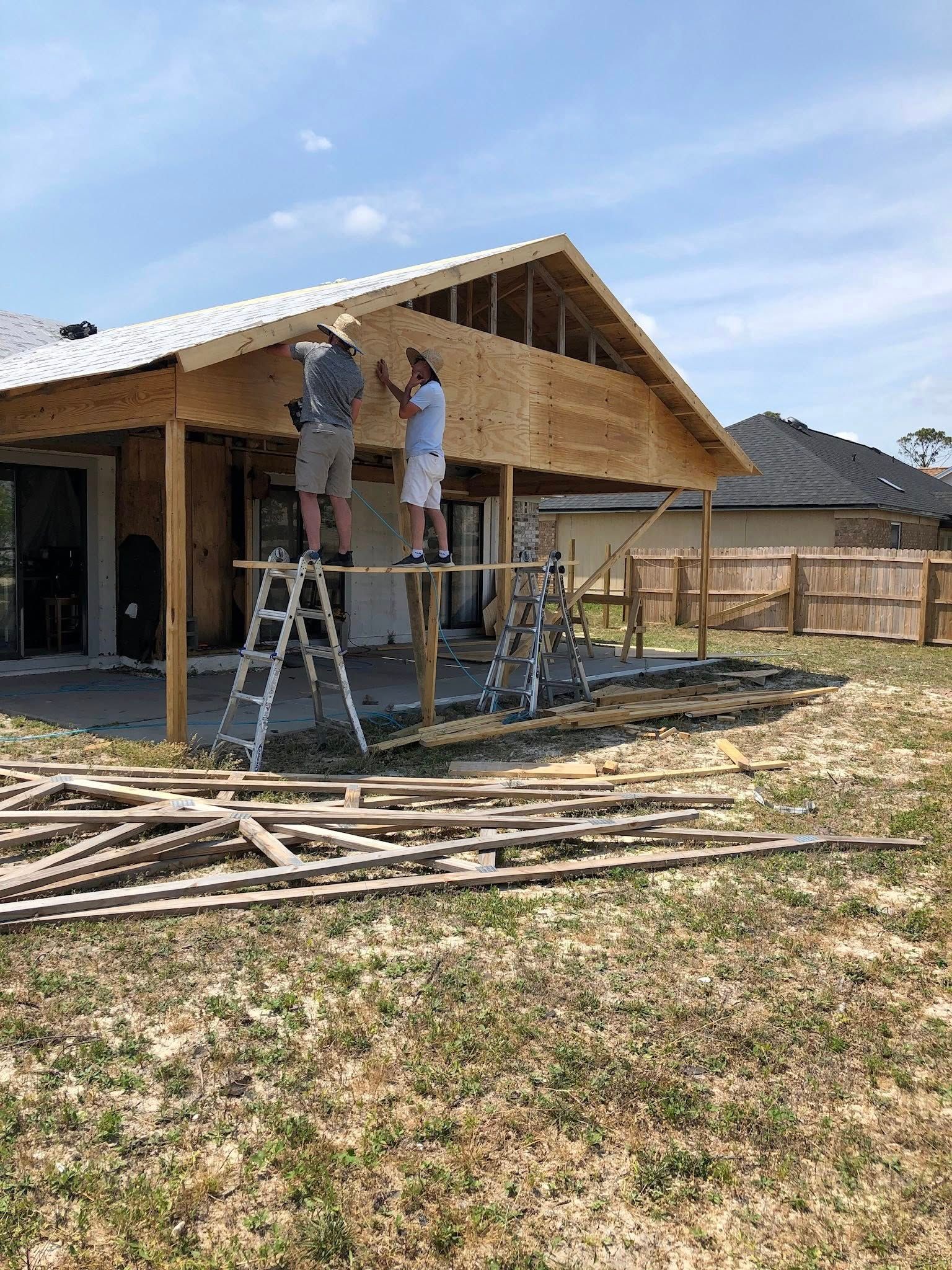 Two workers on ladders install plywood sheeting onto the roof gable of a house under construction.