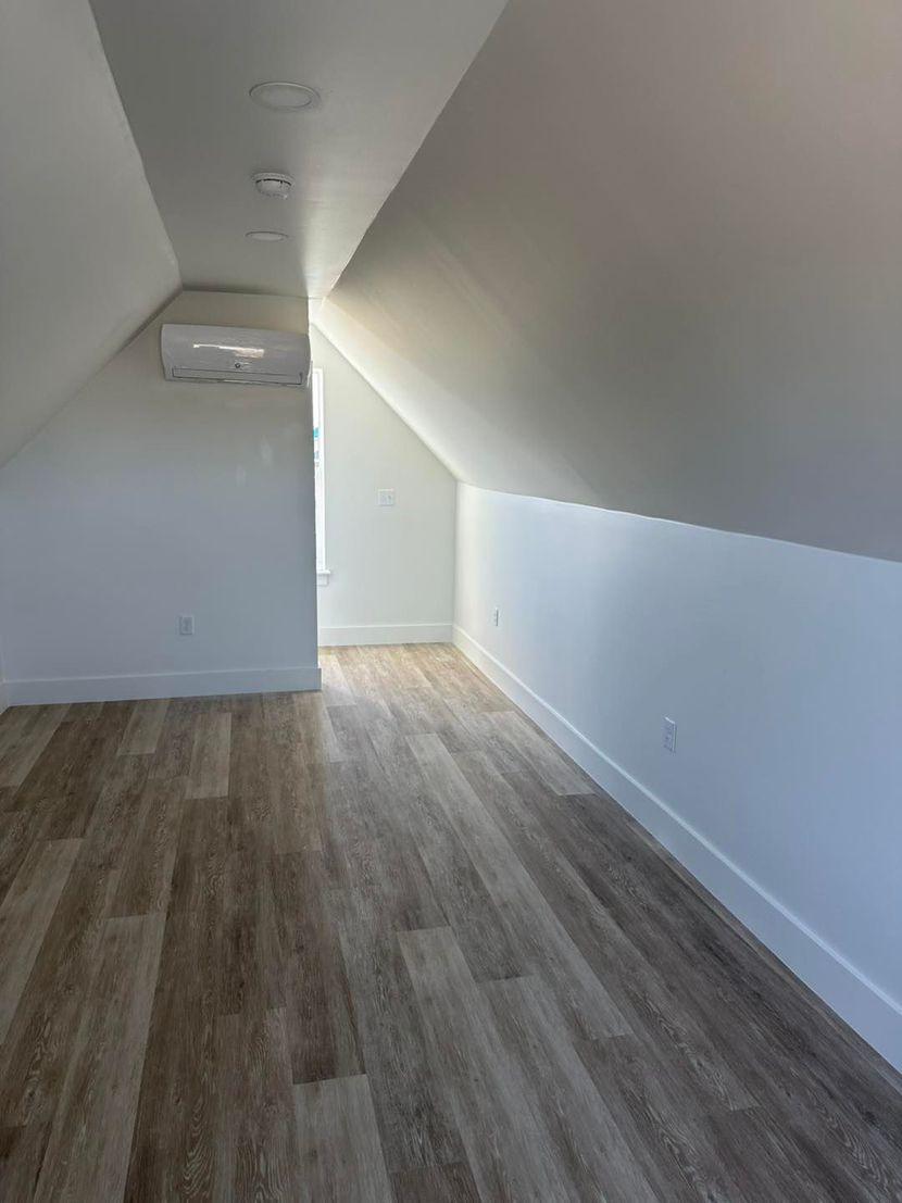 An empty attic room with white slanted walls, a wood-look floor, and a wall-mounted mini-split air conditioner.