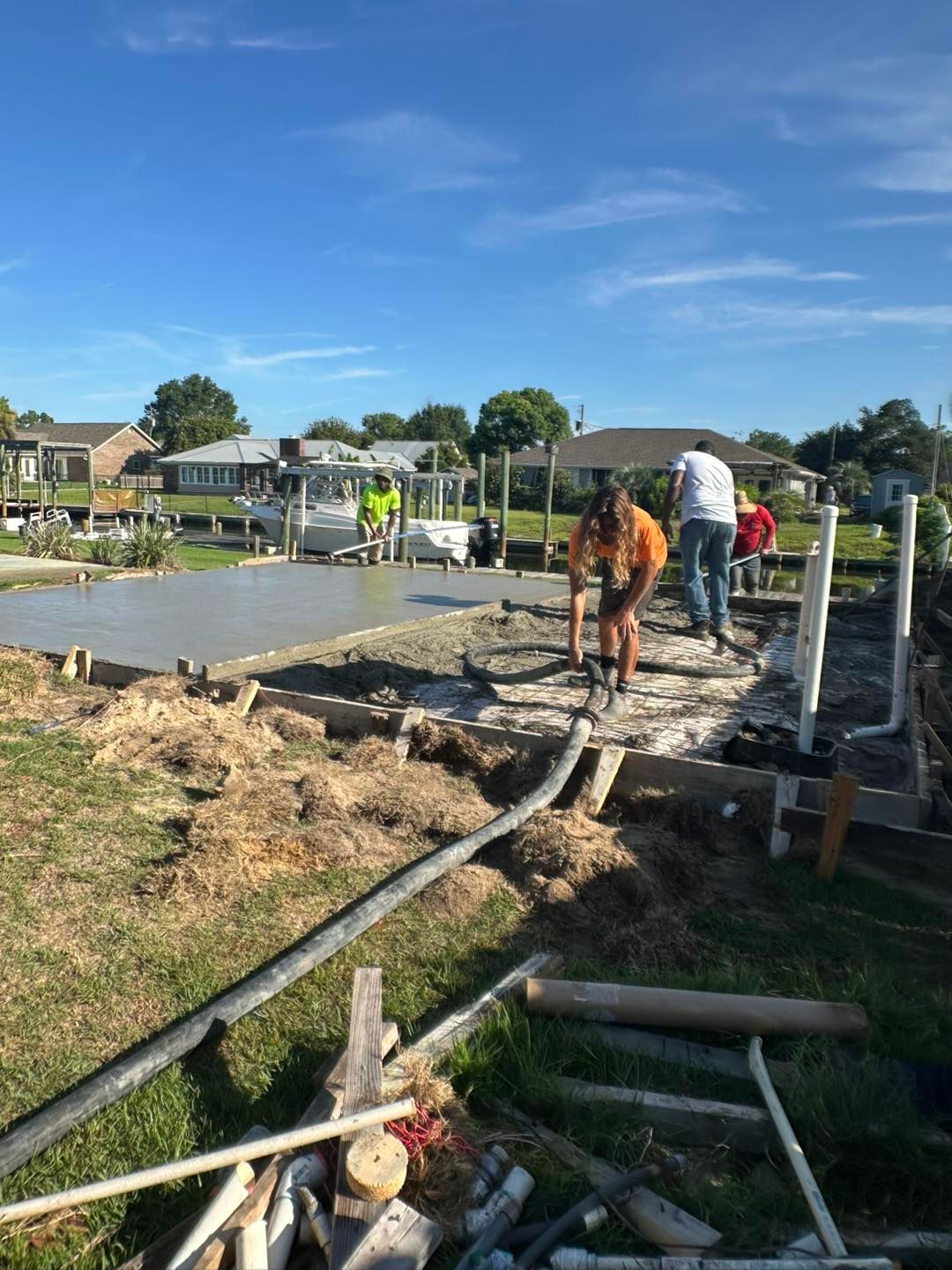 Construction workers pour wet concrete into a wooden frame for a new foundation on a sunny, grassy waterfront lot.