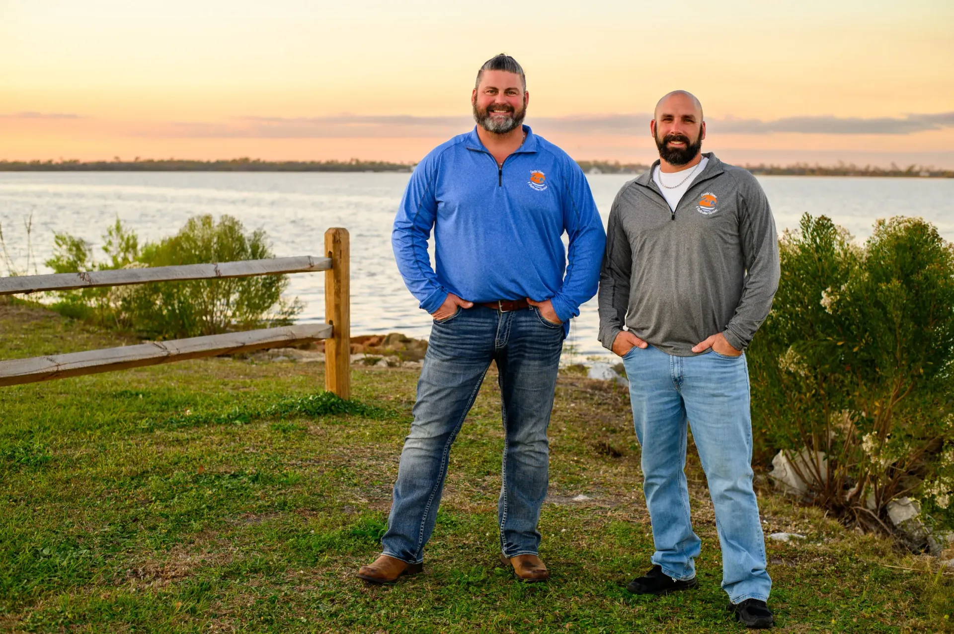 Two men standing side-by-side in casual attire outdoors near a lake at sunset.
