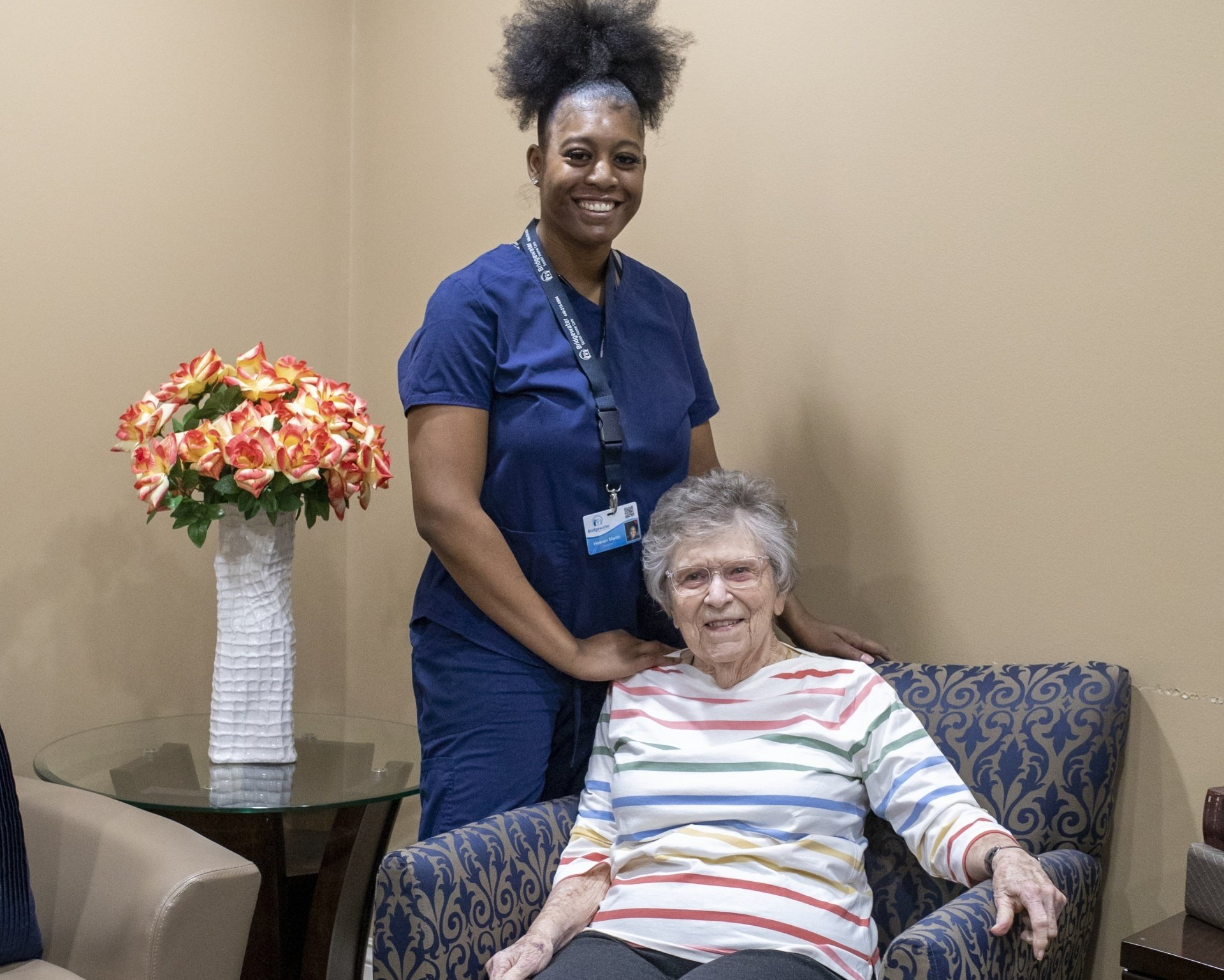 Caregiver smiles, hand on seated woman's shoulder, posing together. A vase of flowers beside them in a room.