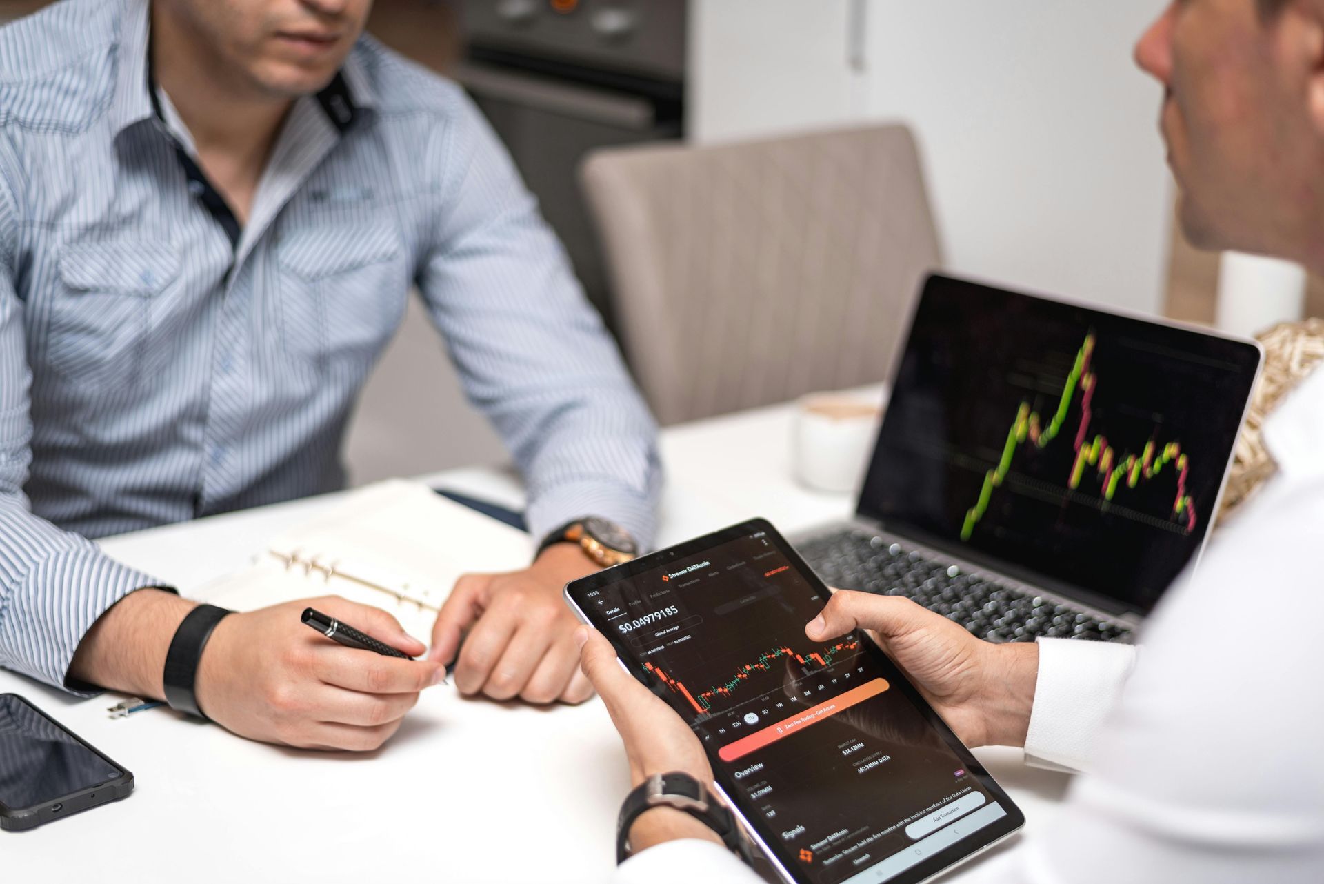 Two people reviewing stock charts on a laptop and smartphone at a desk