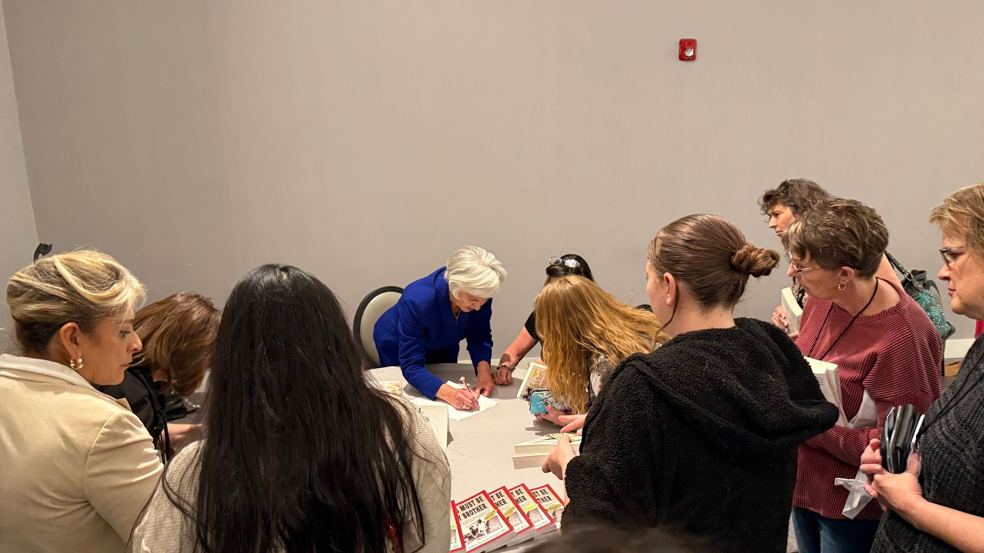 People gathered around a table; a woman in a blue suit signs items.