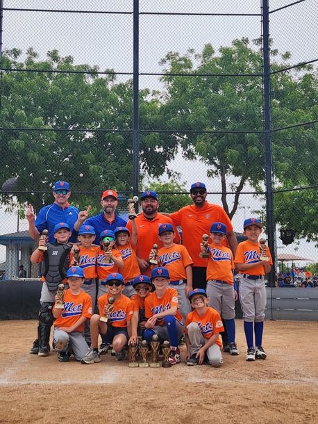Youth baseball team in orange and blue uniforms posing together on a field in front of a chain-link fence