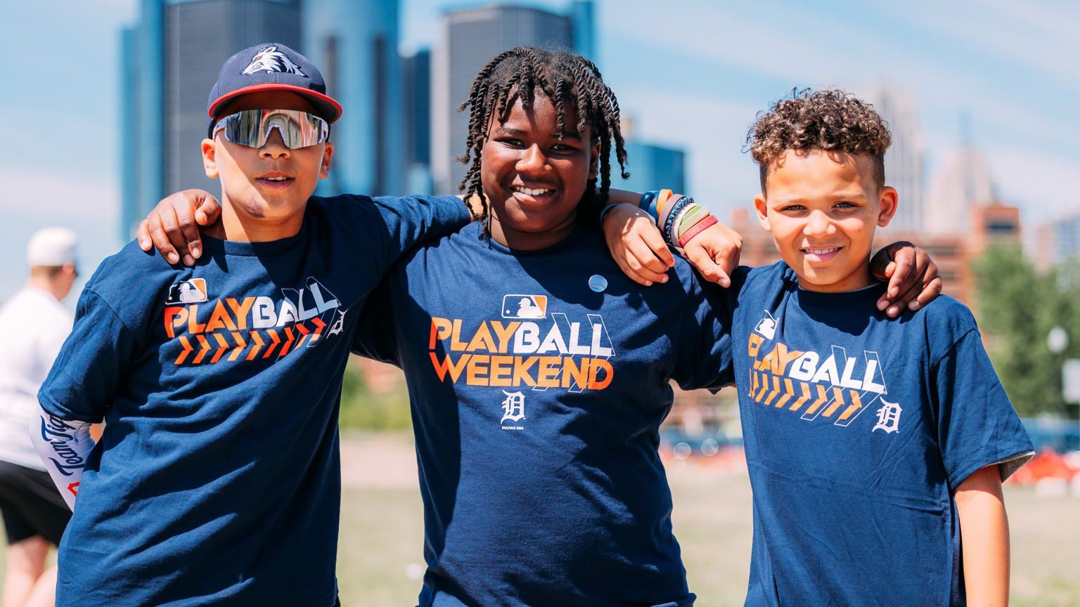 Three smiling teammates in navy “PLAYBALL WEEKEND” shirts standing outdoors with a city skyline behind them