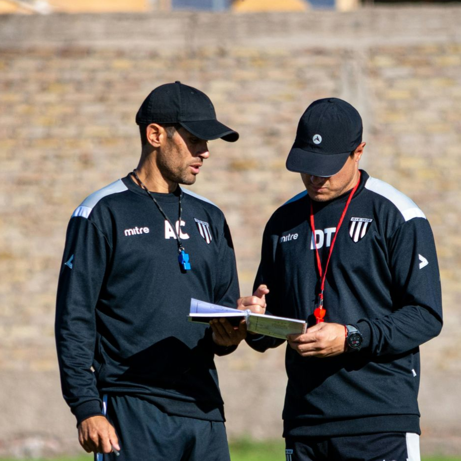 Two coaches in black caps and tracksuits review a clipboard outdoors by a stone wall.